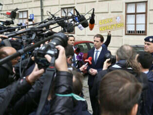 Sebastian Kurz speaks to the media outside the Hofburg Palace, Vienna, as a protester holds a placard reading "Against corruption" in the background