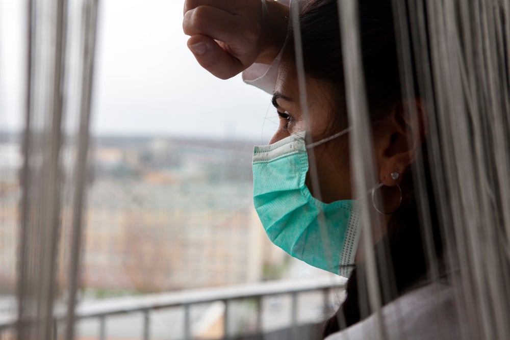 A woman with a medical mask on looking out of her window